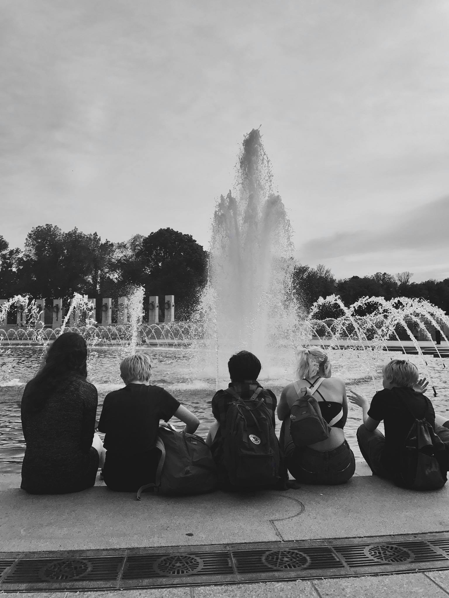 5 youth sitting in front of fountain facing away from camera in black and white
